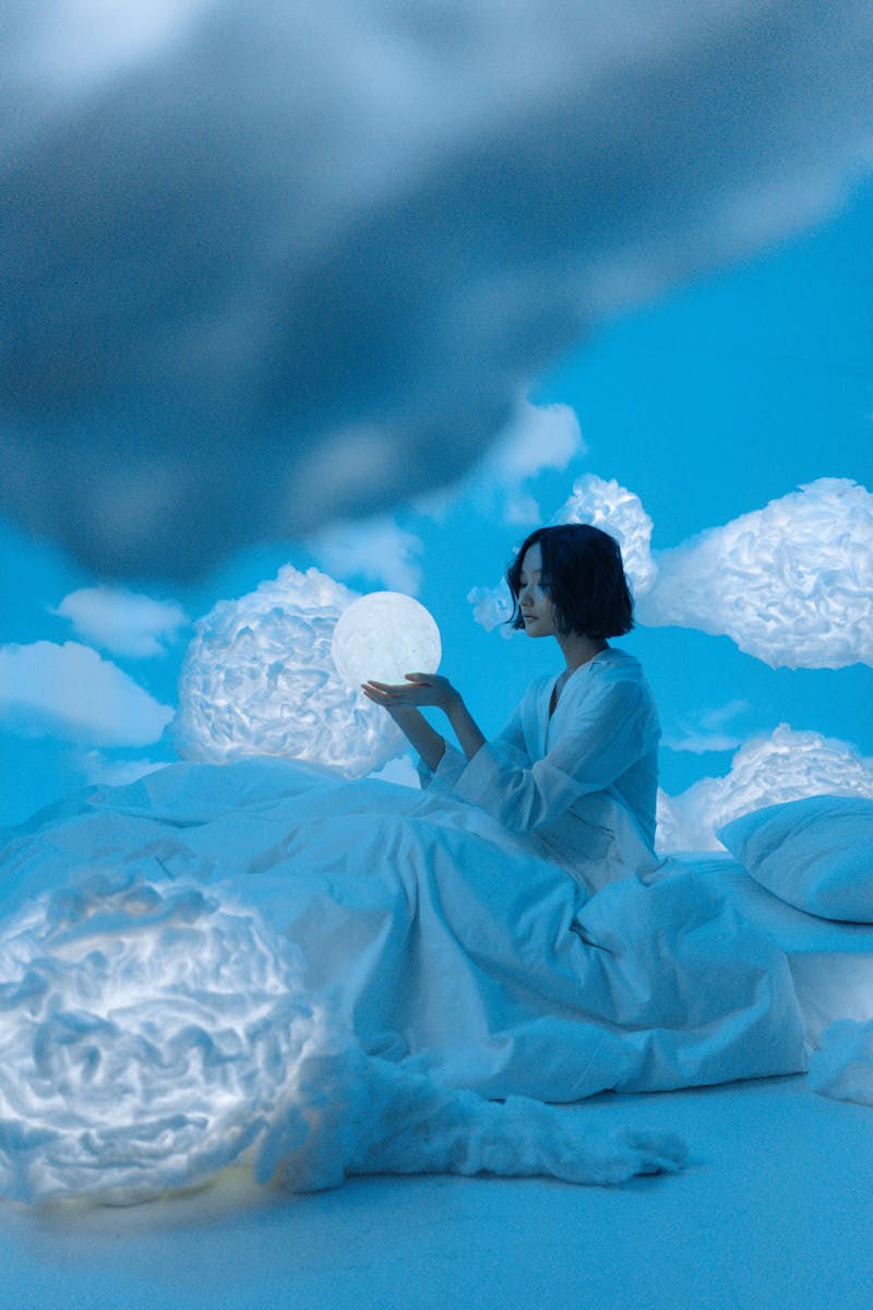 Asian woman holding glowing orb on bed in surreal cloud-filled room, serene night.