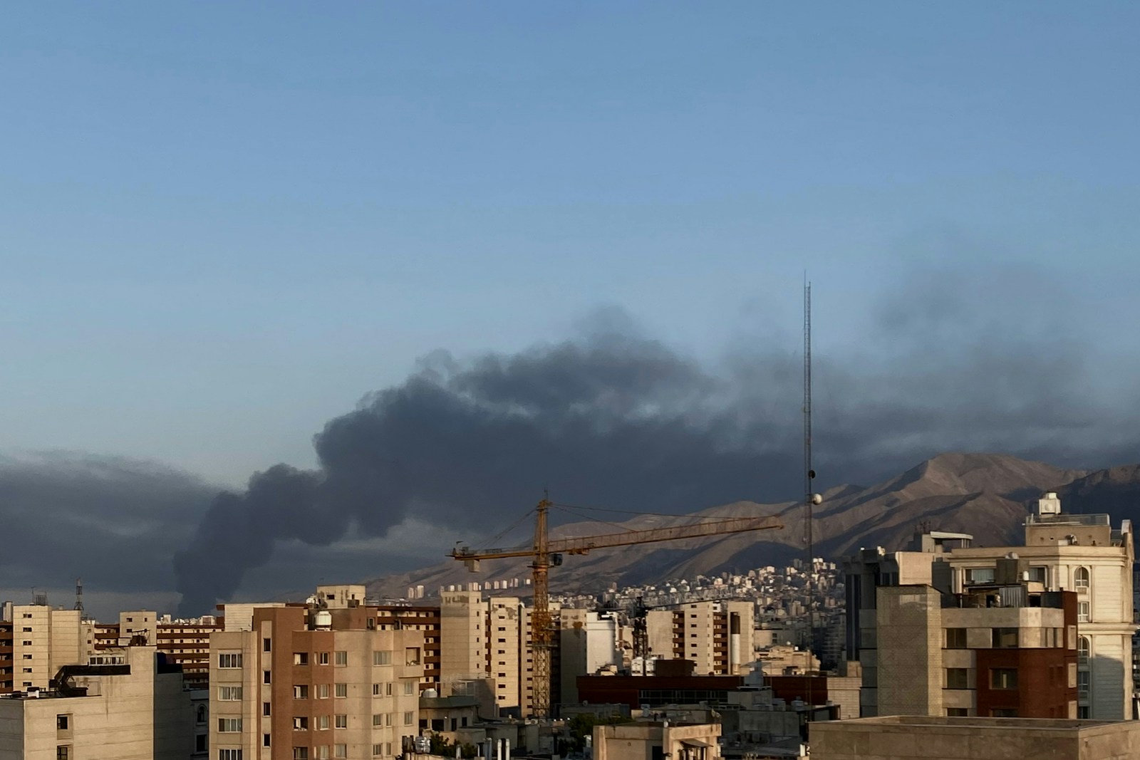 Smoke billows over a cityscape and mountain range.